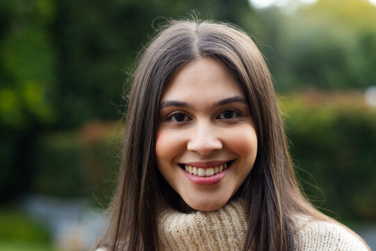 Smiling woman posing in garden while wearing beige knit turtleneck sweater showing blurred hedges