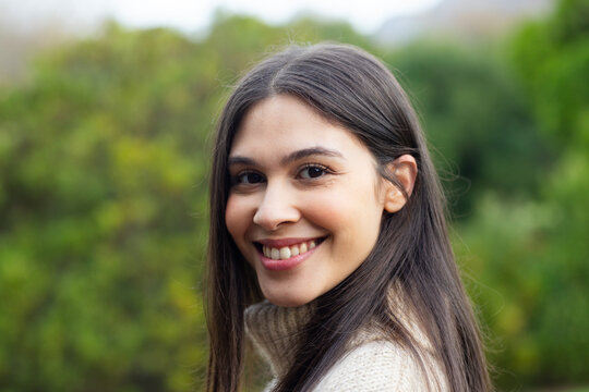 Woman smiling gently wearing cream colored knitted sweater in garden with blurred foliage
