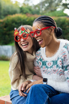 Diverse female friends sitting on stone bench in garden wearing festive glasses laughing together