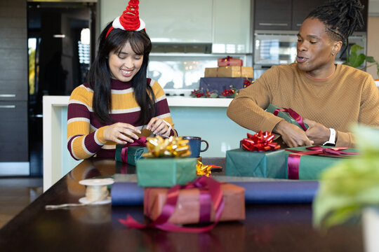 Diverse couple sitting at kitchen table, wrapping Christmas gifts, woman wearing striped sweater