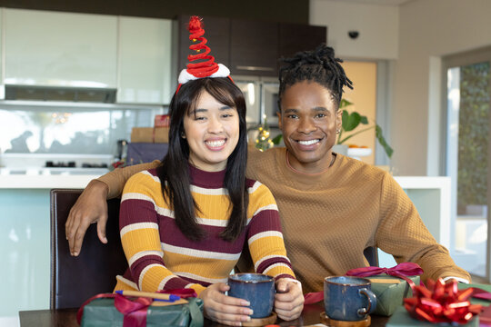 Diverse couple smiling, sitting at dining table in kitchen, wearing Christmas headband
