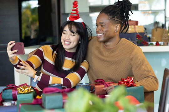 Diverse couple taking selfie with phone in pink case at table, woman wearing Christmas headband