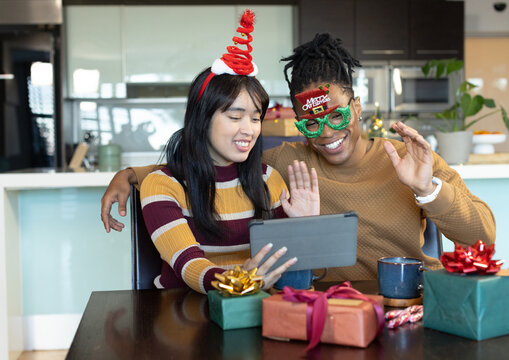Diverse couple waving and smiling while using tablet at kitchen table with wrapped Christmas gifts