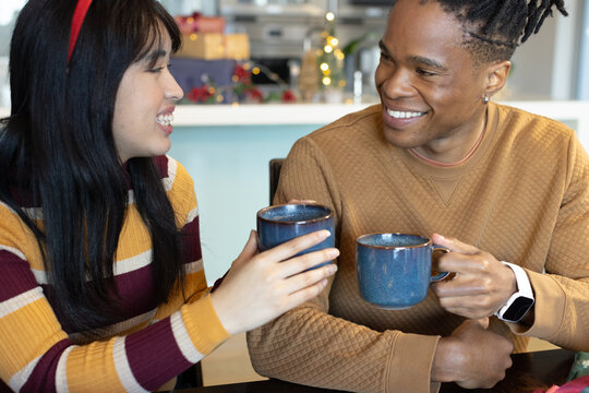 Diverse couple raising blue ceramic mugs at kitchen table with wrapped gifts, celebrating Christmas