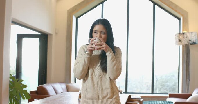 Mid adult woman holding ceramic mug at dining table inhaling aroma savoring warmth in sip