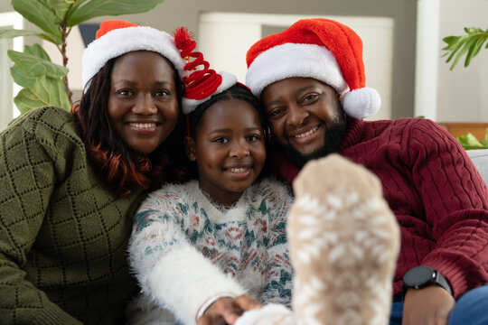 African American family sitting close on sofa wearing Santa hats, Christmas headband, fuzzy slipper