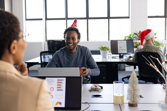 Diverse coworkers exchanging wrapped gift and chatting at office with Christmas tree, Santa hat