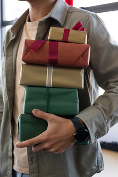 Asian man holding stack of 5 wrapped Christmas gifts by window in khaki shirt showing smartwatch