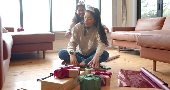 Asian mother wrapping gifts on floor when Indian daughter hugging shoulders affectionately