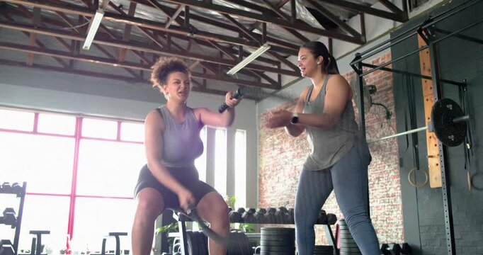 Asian trainer counting on watch urging African-American trainee swinging ropes in gym for intensity