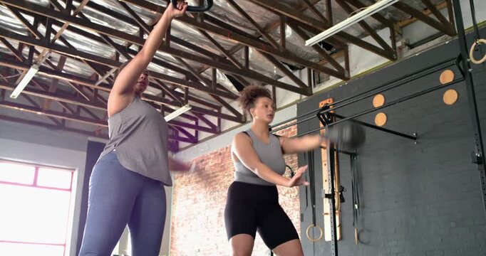 Hispanic and African American women hinging hips, driving kettlebells upward for strength in gym