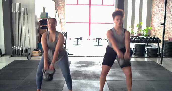 Diverse female friends stepping onto mat in gym initiating kettlebell swinging in sync for strength