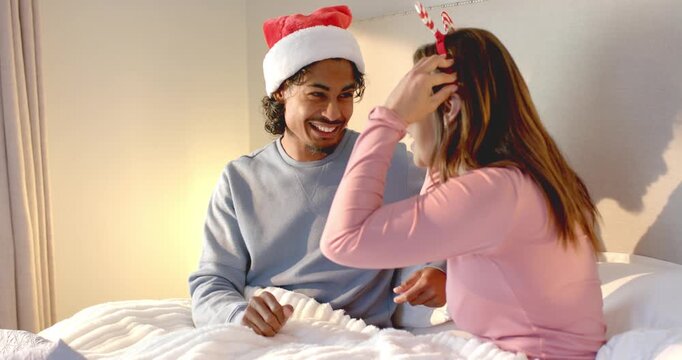 Diverse couple exchanging antler headband laughing capturing moment with smartphone on bed
