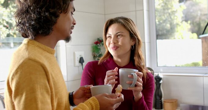 Diverse couple sharing gestures conversing in kitchen by window holding mugs and half-eaten cookie
