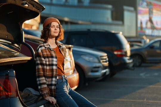 Woman leaning on car trunk in parking lot, beanie and flannel jacket with jeans, smile in warm sunlight; candid lifestyle portrait with authenticity, golden hour glow and emotional storytelling.