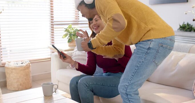 Diverse couple on couch sharing phone screen after grabbing mug, donning hats and sipping coffee