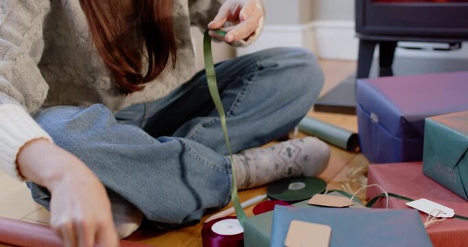 Woman cutting green ribbon, measuring and tying twine, wrapping Christmas gifts by stove
