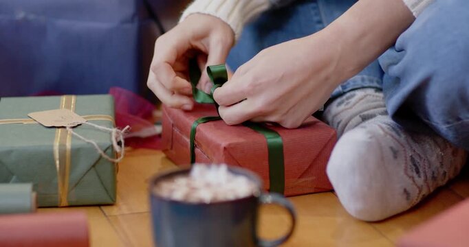 Female tying ribbon bow on red gift in living room, preparing for Christmas with marshmallow mug