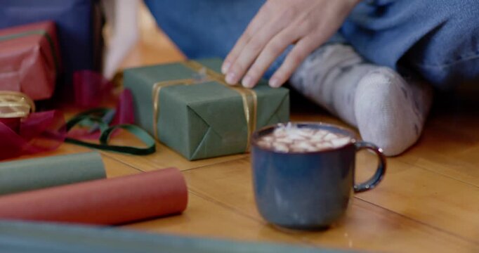 Female tying ribbon on green gift at home, finishing tag then sipping mug for Christmas