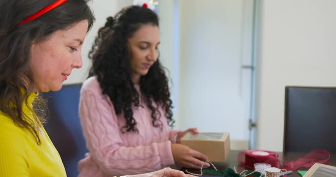 African American women folding boxes, tying green ribbon at table while preparing Christmas gifts
