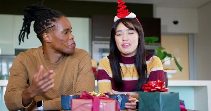 Couple greeting camera, waving, holding wrapped gift and presenting Christmas mugs at kitchen table