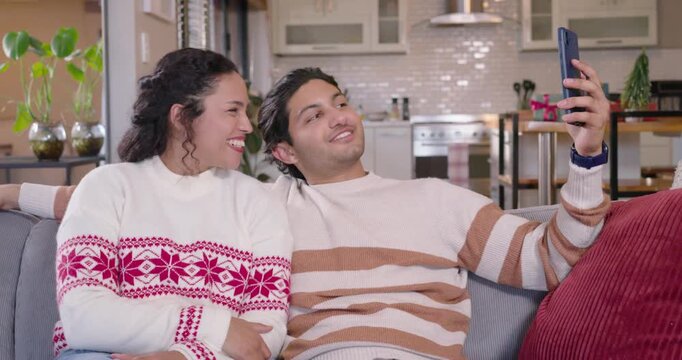 Couple in holiday sweaters using smartphone on couch at home, greeting family during Christmas call