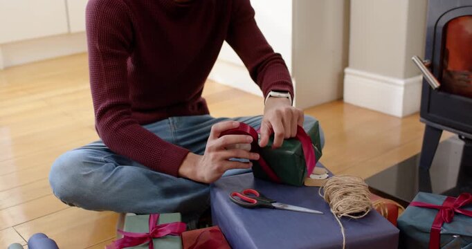 African American man picking up red ribbon spool and scissors, wrapping Christmas gifts on floor
