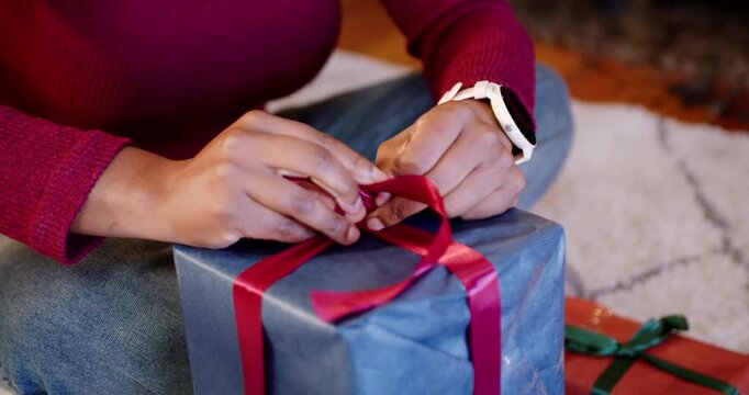 African American woman starting with ribbon across blue box, tying red bow on rug for Christmas