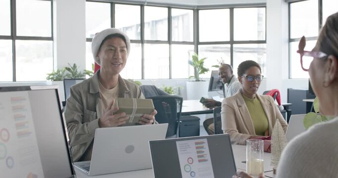 Diverse coworkers at shared desk, Asian man in Santa hat handing wrapped gift celebrating Christmas