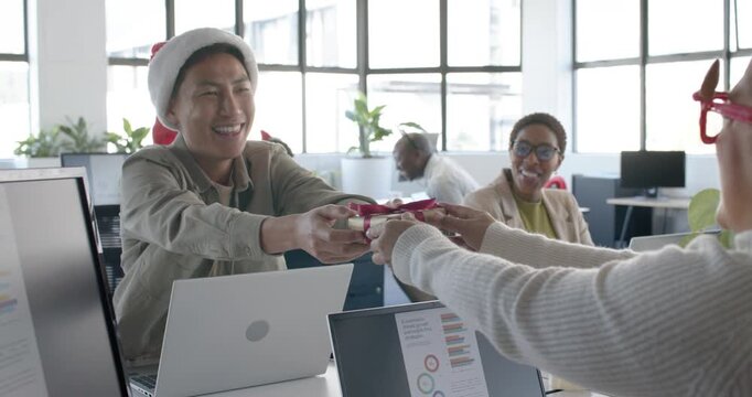 Diverse team sharing Christmas gifts at office, woman extending box, Asian man in Santa hat opening