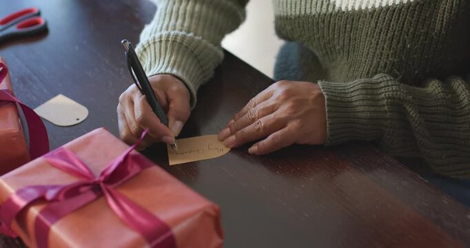 Woman writing name on brown tag with black pen at wooden table preparing Christmas gifts