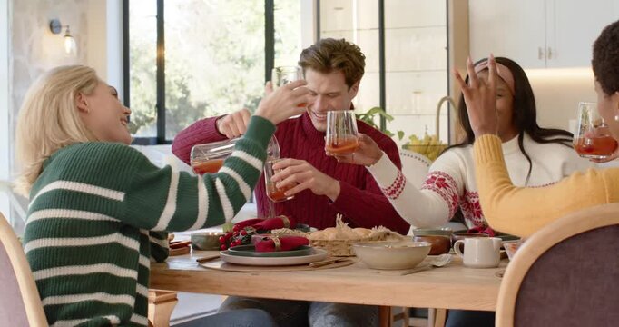 Diverse friends in festive knits pouring pitcher into glasses, clinking at table for Christmas