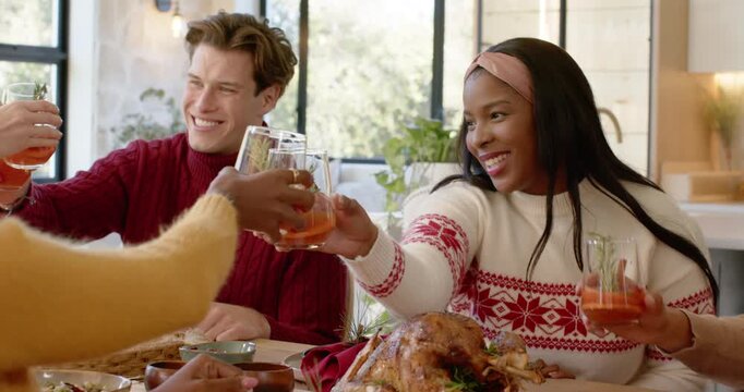 Diverse friends smiling, reaching and clinking stemless glasses above turkey at table for Christmas