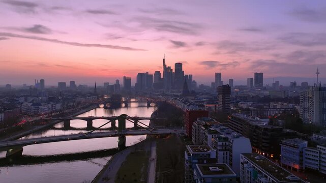 Aerial view of Flosserbrucke spanning the Main River with the illuminated Frankfurt skyline in the background during a vibrant sunset in Frankfurt am Main, Hessen, Germany.