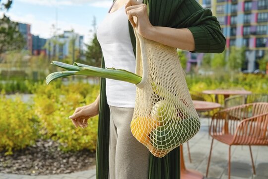 Close-up view of string bag with fresh vegetables and fruits on shoulder of woman walking through residential area. Concept of healthy lifestyle and reusable household items