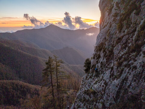 Aerial view of a climber scaling a steep rock face in the Apuan Alps at sunset with rolling mountain ridges in the background in Minucciano, Tuscany, Italy.