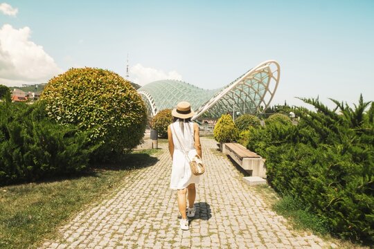 Woman in white dress and straw hat walking toward Bridge of Peace. Rike Park landscape, modern architecture, summer travel, sunny city sightseeing tour. Tbilisi. Georgia