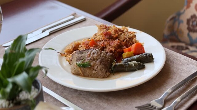 Close-up of a plated Middle Eastern meal featuring rice, meat, carrots, and stuffed grape leaves. The delicious lunch sits on a restaurant dining table with silverware and a small potted plant.