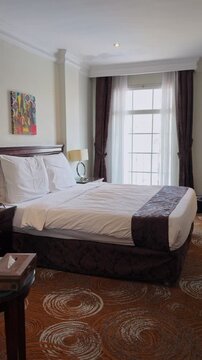 Vertical view of an empty hotel bedroom interior during daytime. A tidy bed with white linens and a brown runner sits on a patterned carpet next to a bright window with sheer curtains.