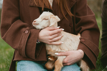 Gentle scene of a woman cuddling a soft goat kid outdoors on a grassy field, embodying warmth, care, and rural lifestyle moments with a calm, intimate mood. It captures a caring moment between human © SHOTPRIME STUDIO