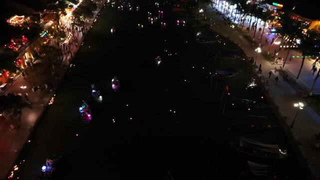 Colorful boats with bright lights move smoothly on a dark river in Hoi An, Vietnam, with lively street festivities and reflections visible nearby