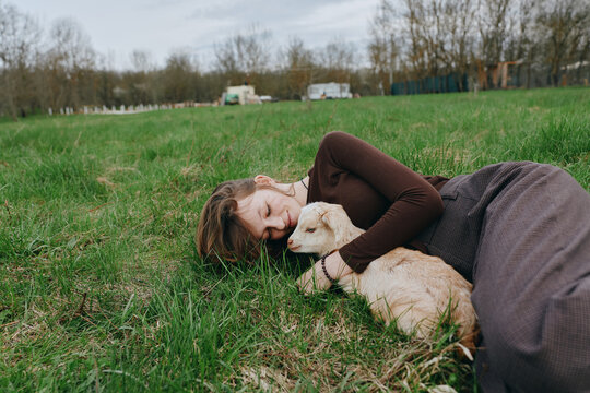 Young woman lying on grass cuddles a small goat in a peaceful outdoor setting during early spring, showcasing a calm and tender lifestyle moment.
