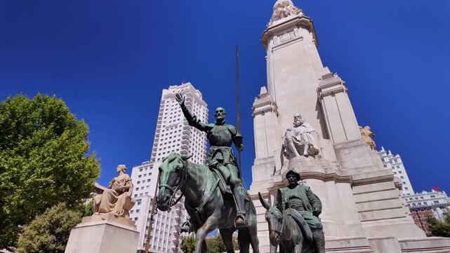 Wide low angle shot of the Cervantes Monument with Don Quixote and Sancho Panza statues under a deep blue Madrid sky.