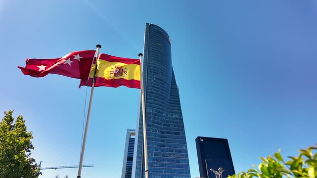 Madrid and Spanish flags waving beside Torre de Cristal in the Cuatro Torres business district under a clear blue sky.