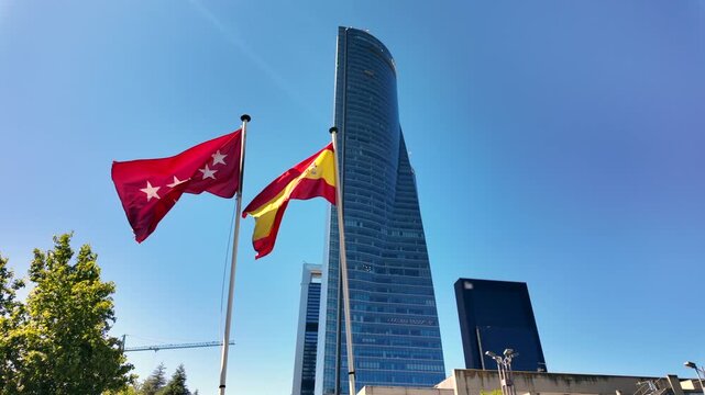 Wide low angle view of Spanish and Madrid flags with the Torre de Cristal skyscraper and clear blue sky behind them.