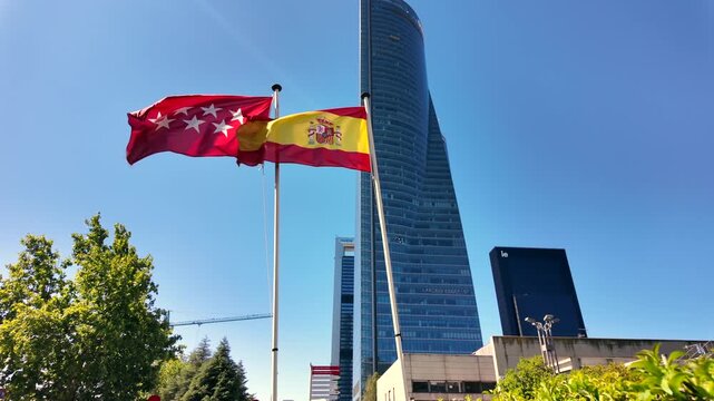 Spanish and Madrid regional flags waving on flagpoles with Torre de Cristal rising behind them in the Cuatro Torres area.
