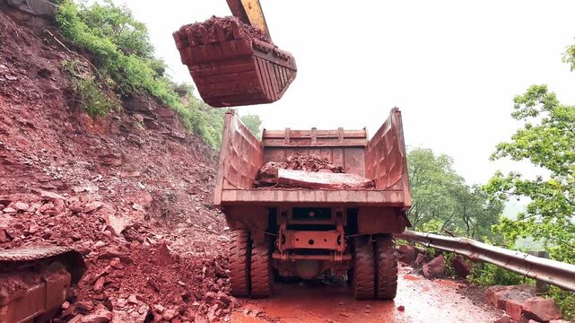 Static shot of an excavator loading reddish soil and rocks into a dumper truck during landslide debris clearance on a mountain highway