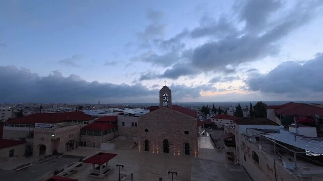 Stone bell tower crowns St George Greek Orthodox Church at dusk in Madaba, Jordan, red tile rooftops spread across hillside town beneath dramatic gathering storm clouds. Aerial drone timelapse