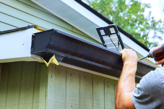 Contractor attaches gutter to edge of building while using drill with house with green siding.