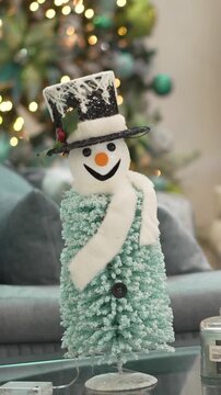 A snowman sits at a table in the decorated living room in front of a Christmas tree. 4k.